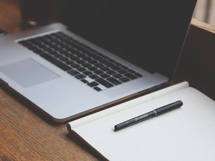 Foto eines Laptops. Daneben liegt ein Schreibblock und ein Stift darauf.